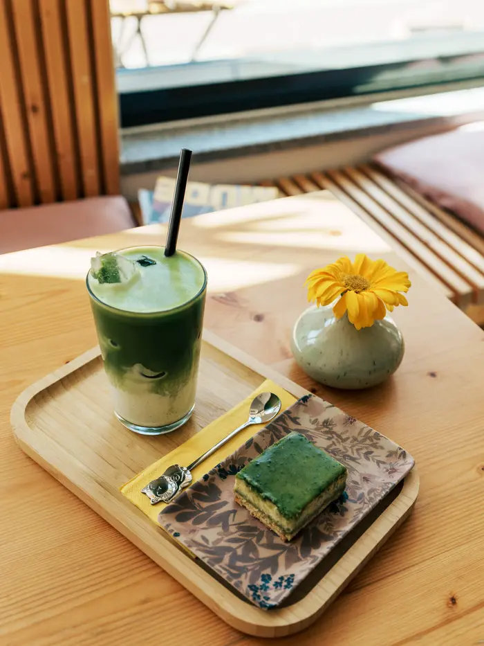 Iced Matcha und ein Stück Matcha-Kuchen auf einem Tablett mit kleiner Blumenvase.
